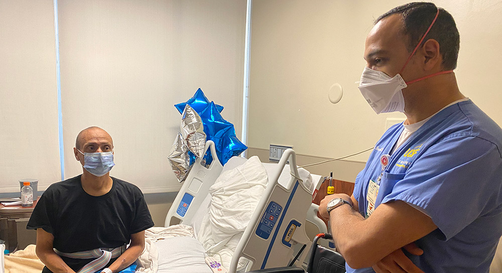 A male patient sits on a hospital bed next to a healthcare worker in a mask