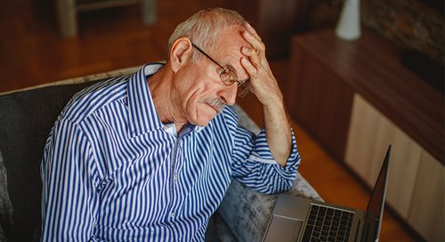 20220920-behavioral-health A senior citizen, white and male, rests his head in his hands and looks forlorn. He is wearing a blue and white striped shirt and looking at a laptop computer.