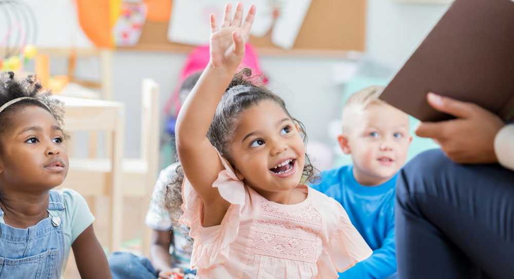 A little girl sits on a mat with her classmates and raises her hand