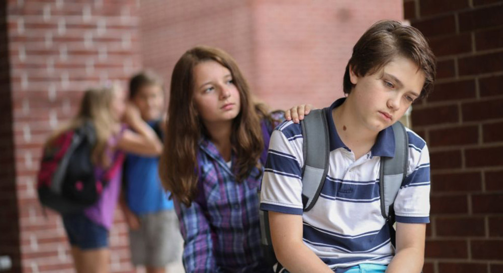 a young teen girl puts her hand on the shoulder of a sad, young teen boy