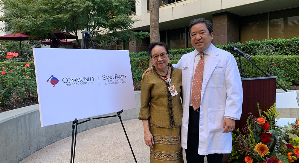 A man and a woman stand next to a sign reading "Community Medical Centers | Sang Family Postpartum & Newborn Care"