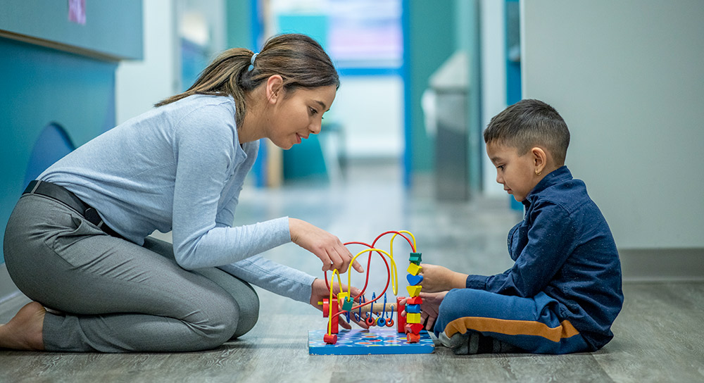 A young Latina sits on the floor opposite a Latino toddler. In between them is a colorful toy that they both are invested in.