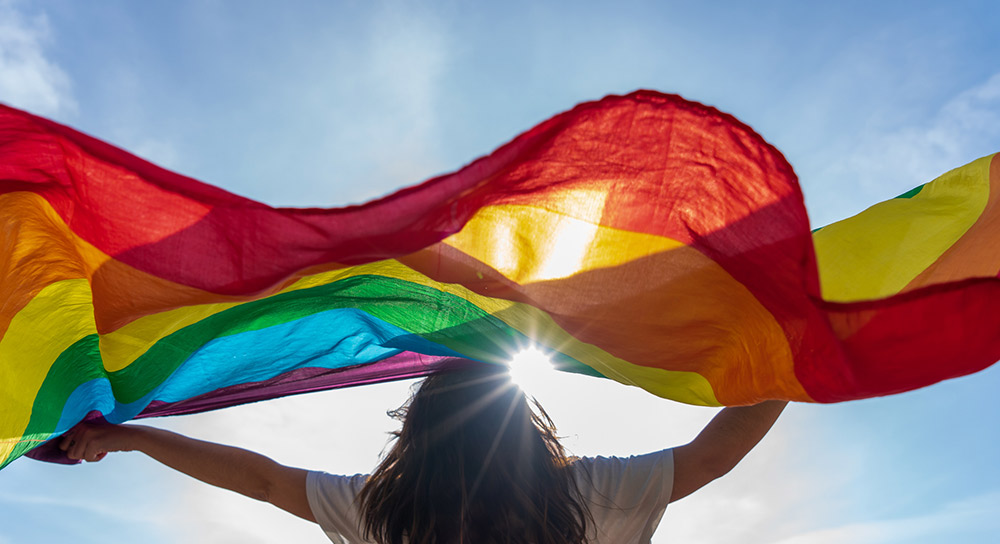 person outside in the shining sun holding a rainbow pride flag