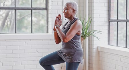 A woman in her 50s who is Black stands with her hands pressed together and one knee raised in a yoga position.