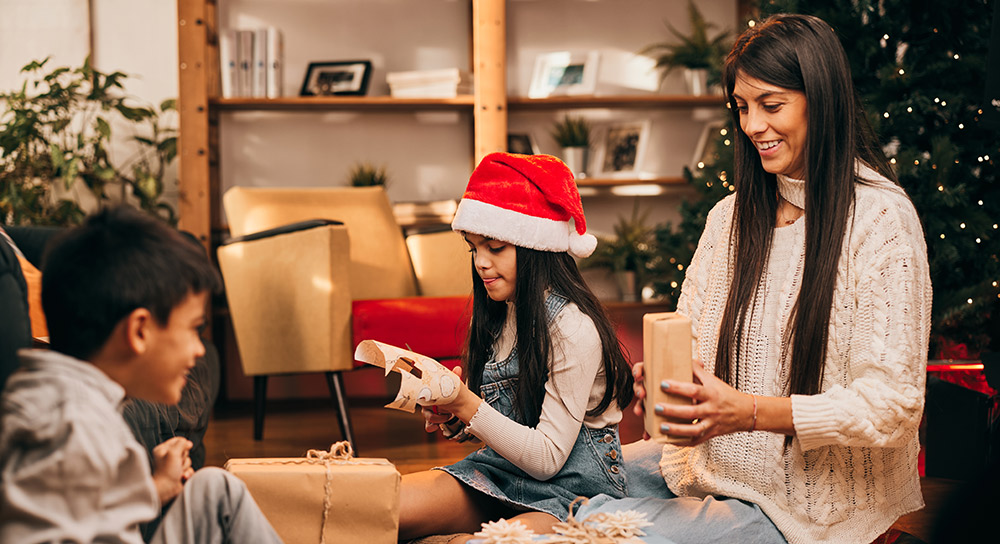 A mother and her two children -- a boy and a girl -- sit in front of their Christmas tree wrapping gifts.