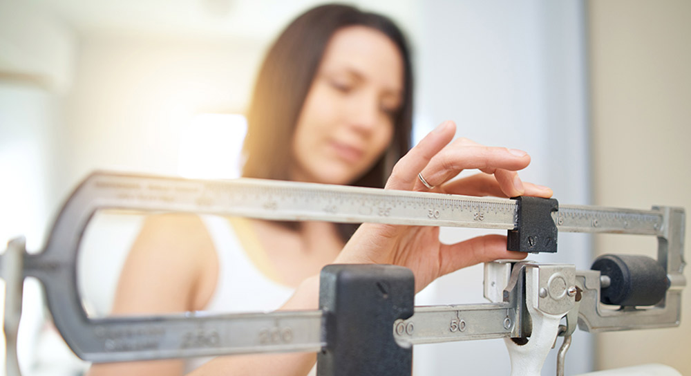 A scale in the foreground, behind it, a woman moves the weights