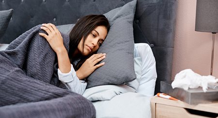 As young woman with dark brown hair and light skin lays in bed looking miserable from sickness.