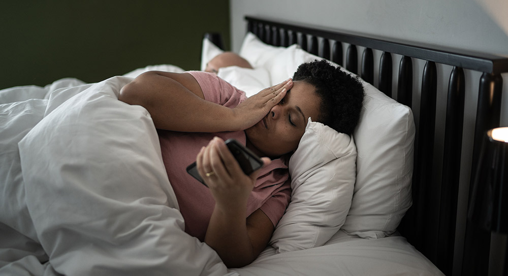 A woman holds her head and looks at her phone in bed