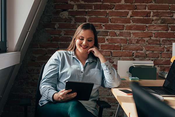 A young woman in a public workspace looks at her work tablet and smiles