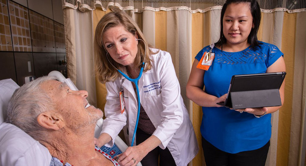 A white older man lies in a hospital bed while a white female doctor uses a stethoscope to hear his heart. A younger, Asian woman stands next to the doctor with a tablet. 