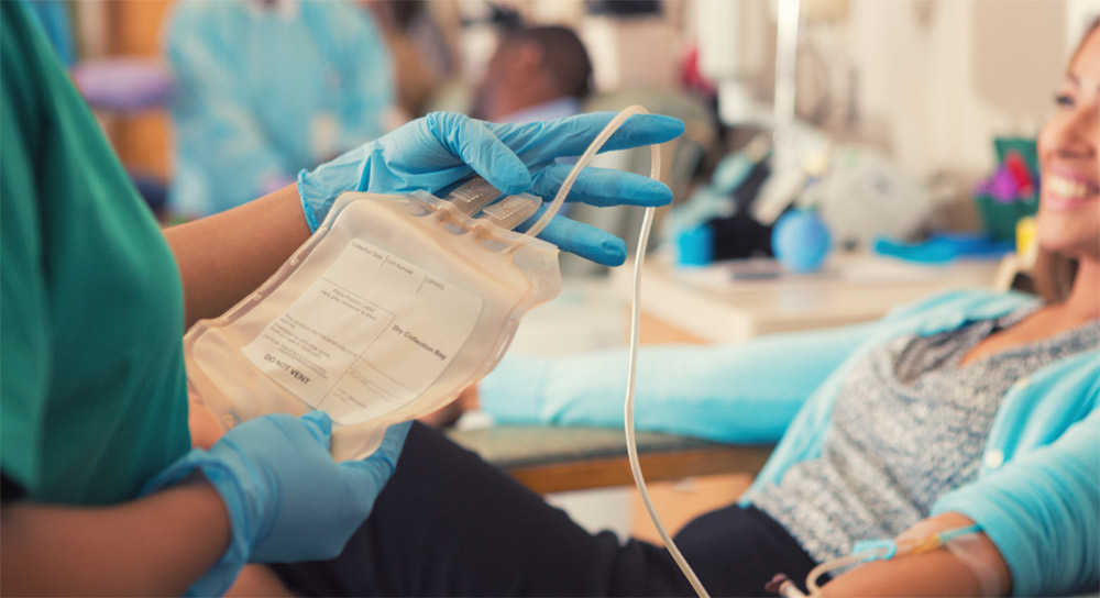 Close up of an empty blood bag held by gloved hands. In the background, a smiling young Asian woman sits in a chair ready to donate blood.