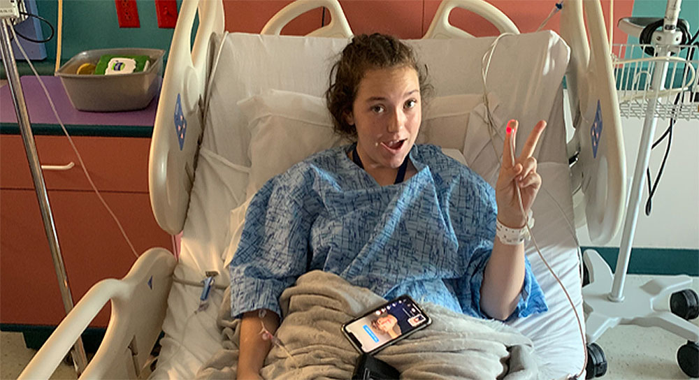Young woman in hospital bed holds up her fingers in a "peace" sign