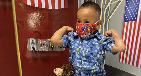 A young boy wearing a face mask and a Hawaiian shirt flexes his biceps.