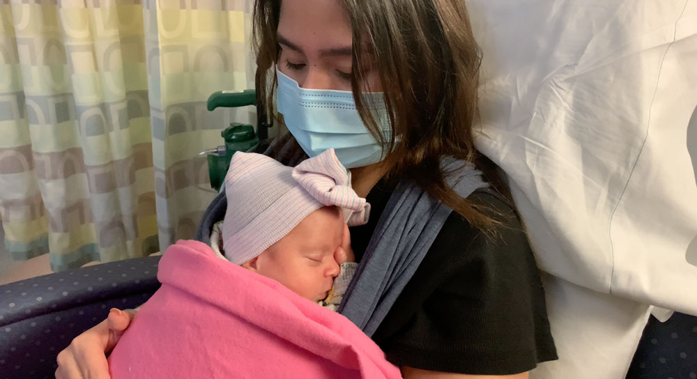Young mother Gabby Manoz, masked and sitting in a hospital chair, holds her newborn to her chest.